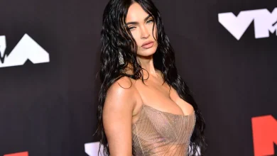 Megan Fox in cropped top & pleated skirt, promoting "Overcompensating" with empowering message.
