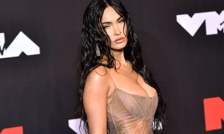 Megan Fox in cropped top & pleated skirt, promoting "Overcompensating" with empowering message.
